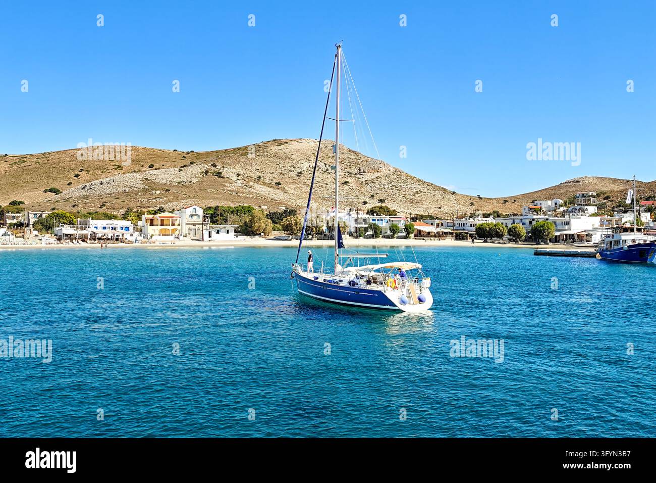 The picturesque harbor bay of the Greek island of Pserimos in the Aegean Sea. A modern blue-and-white sailing yacht lies peacefully in the crystal-clear, turquoise-blue water. In the background stretches the fine sandy beach of the island village, lined with the typical whitewashed houses and inviting taverns - Smartphone Captured Stock Image