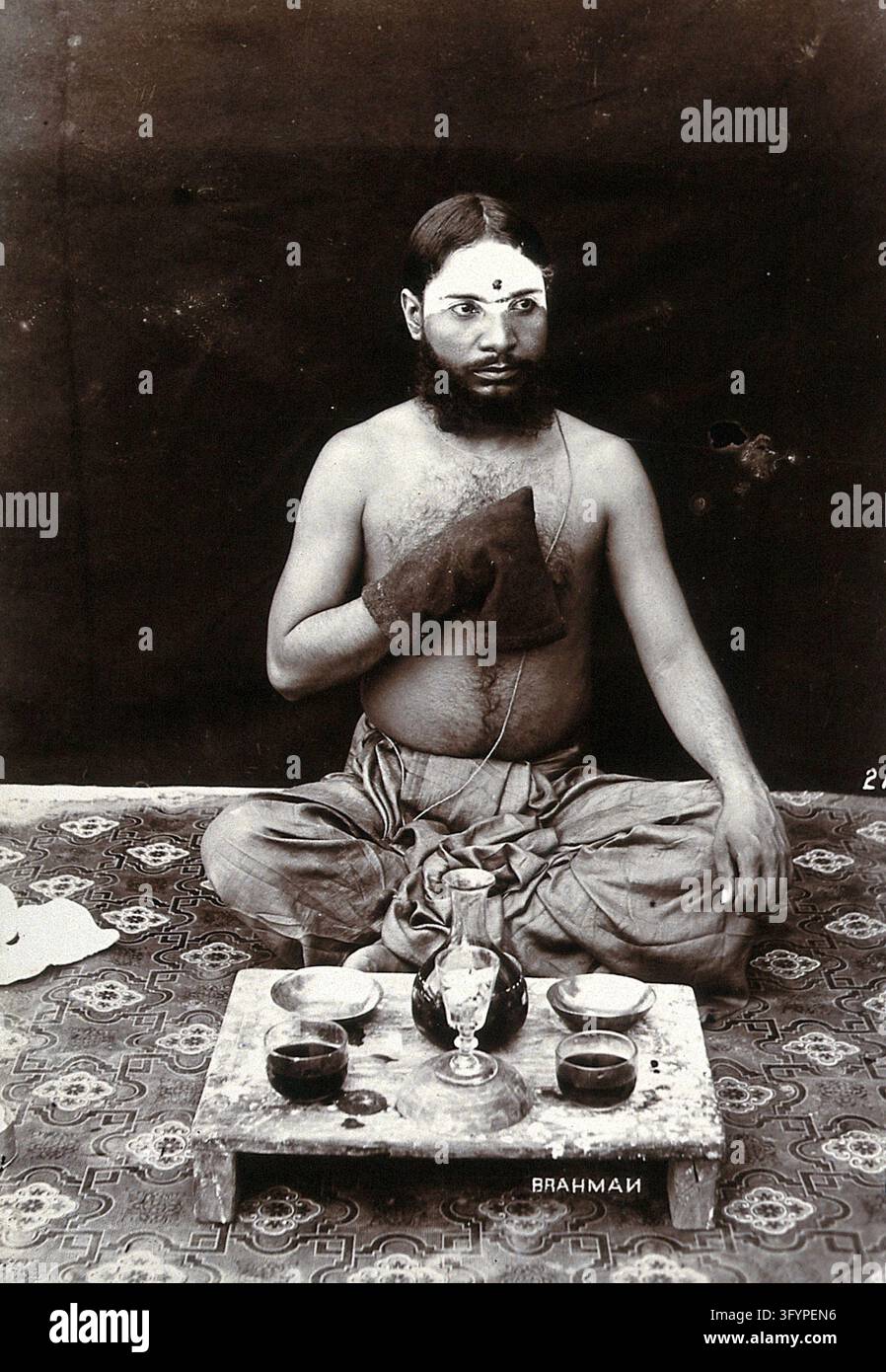 An Indian man wearing a Parsi string and make-up, squatting on a rug ...