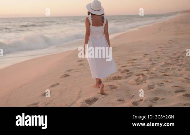 A carefree woman in a flowing white dress and hat walks along a sandy ...