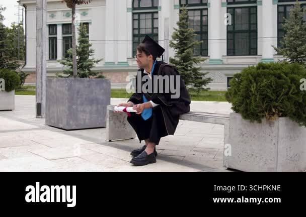 Happy graduate in graduation clothes, black gown and cap, sitting on a ...