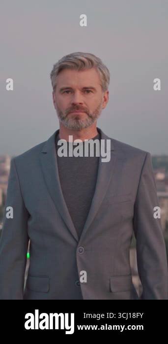 Serious young business man in suit standing look at camera on roof top ...
