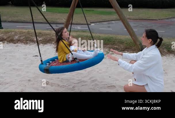 Side view of a mother pushing a round net swing carrying an older girl ...
