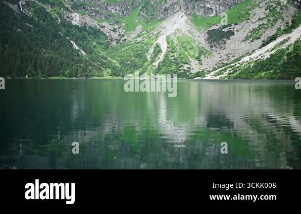 Mountain lake with crystal clear water reflects green slopes and rocky ...