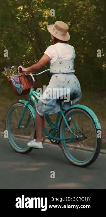 Stylish brunette woman in summer dress and straw hat is riding on retro ...