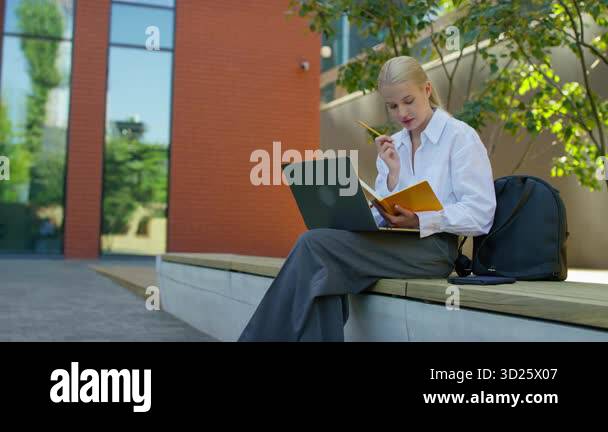 Young woman sitting on a bench in a modern courtyard, writing in a ...