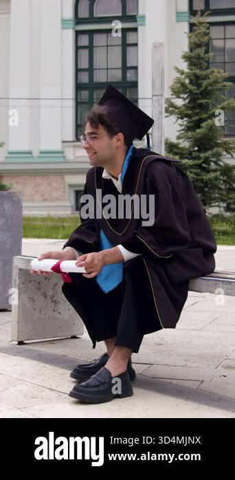 Happy graduate in graduation clothes, black gown and cap, sitting on a ...