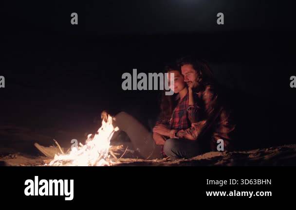 A romantic couple sits by a glowing campfire on the beach at night ...