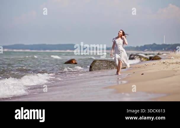 Young woman in white dress walks barefoot along sandy Baltic Sea Beach ...