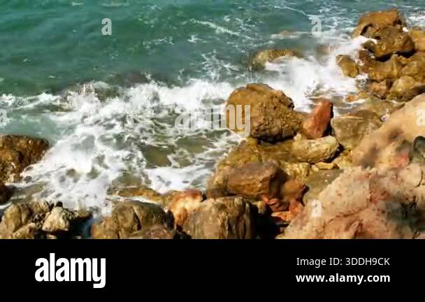 Slow-motion emerald waves crash against sunlit coastal rocks, bursting ...