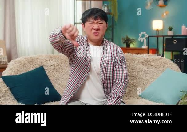 Asian man shows thumbs down gesture frowning unhappily to express ...