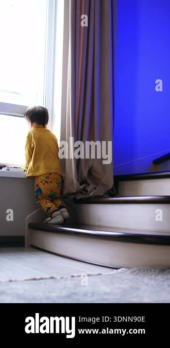 Rear view of a baby boy in yellow clothes standing near the window. Kid ...