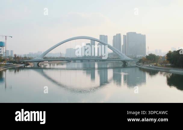 Modern arch bridge spanning calm river with contemporary city skyline ...