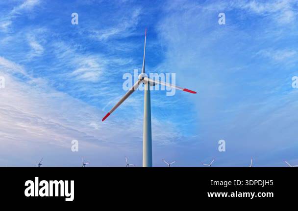 A tall wind turbine is set in a wind farm. The sky is blue with white ...