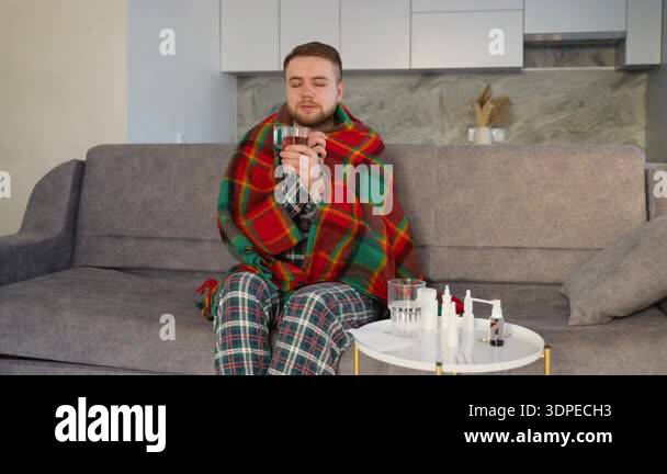 Young man with a cold or flu sitting on a couch, wrapped in a plaid ...