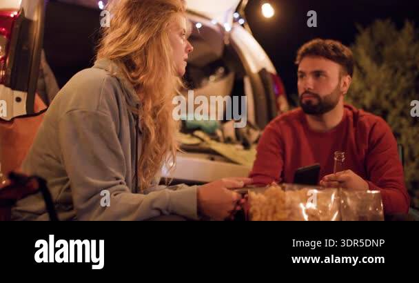 Two people sit by their car at night. They share snacks from a bag and ...