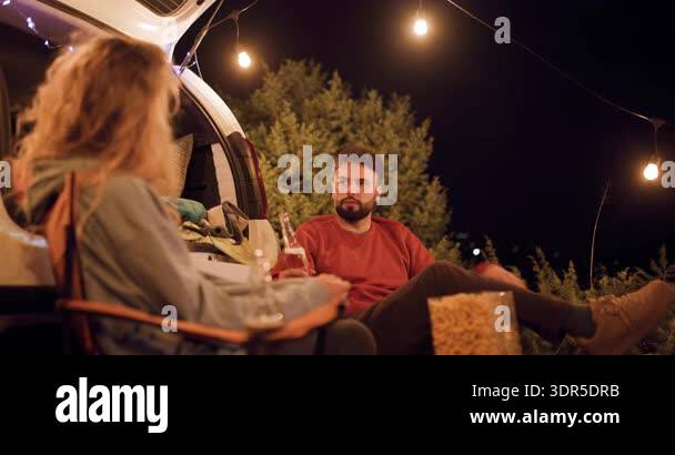 Two friends sit together near a van at night. They talk and laugh while ...