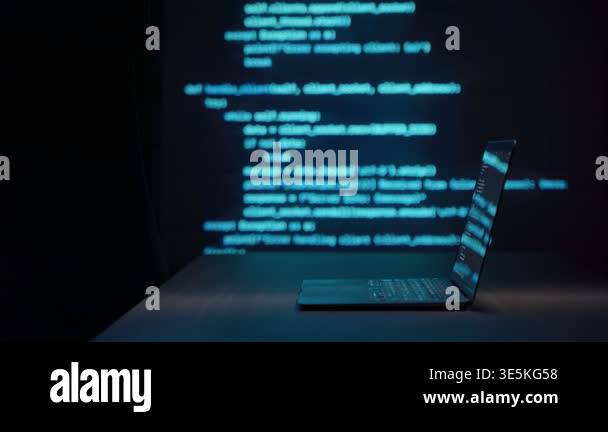 Laptop on dark desk with blue programming code projected in background ...