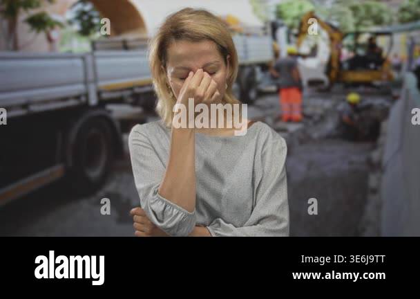 Woman pinches nose with closed eyes beside excavator and truck amid ...