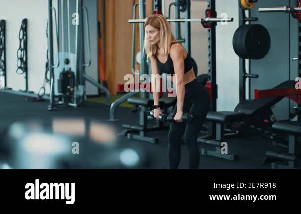 A woman with light skin is lifting a dumbbell in a gym. She focuses on ...