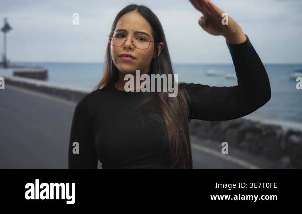 Young latin woman saluting on a seaside promenade with ocean backdrop ...