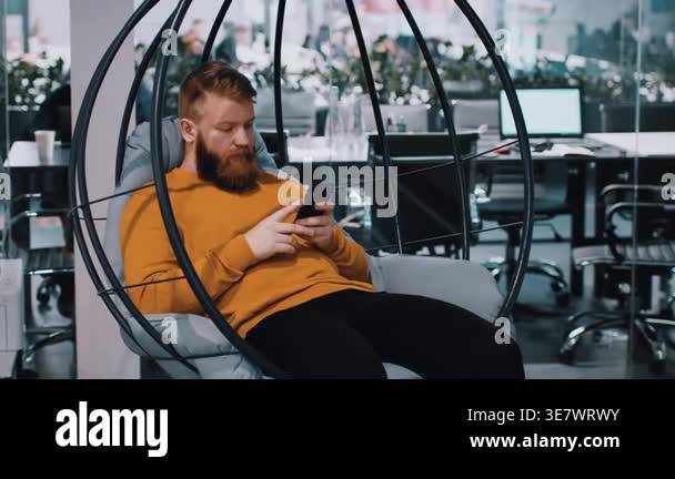 A man with a beard sits on a hanging chair while using a smartphone in ...