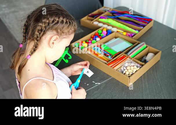Little girl cutting paper with scissors while making craft at table ...