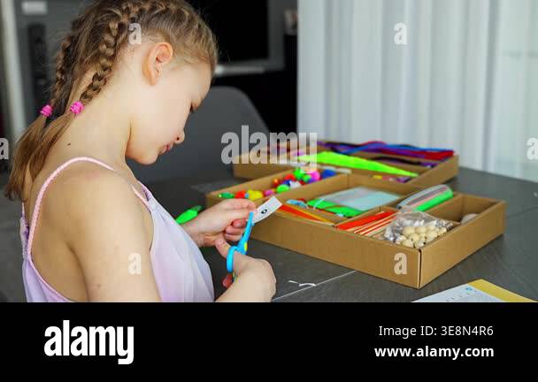 Little girl cutting paper with scissors while making craft at table ...