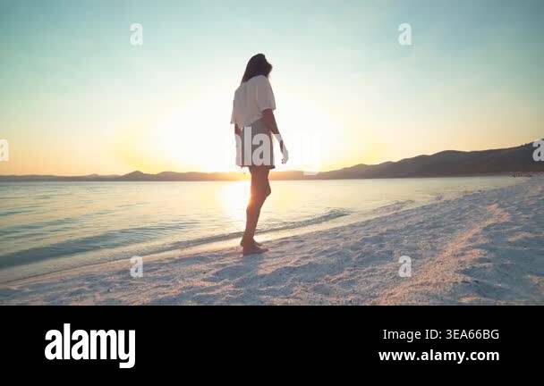 Woman relaxes and walks along white sand beach at sunset beside calm ...