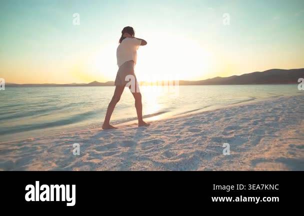 Woman relaxes and walks along white sand beach at sunset beside calm ...