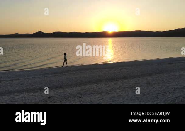 Woman relaxes and walks along white sand beach at sunset beside calm ...