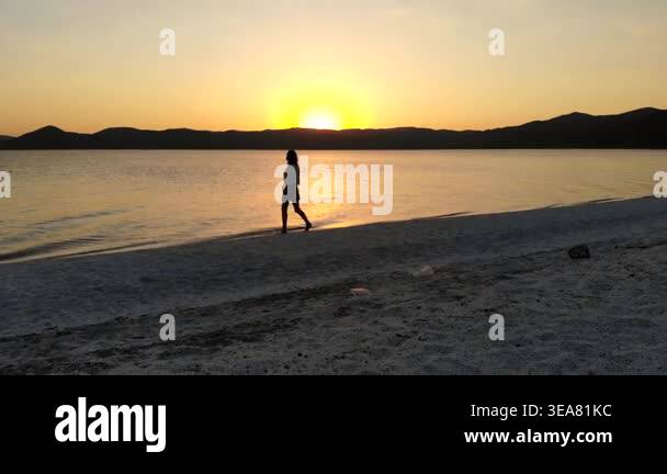 Woman relaxes and walks along white sand beach at sunset beside calm ...