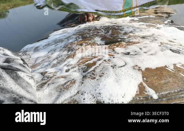 Washing Clothes Using Rocks A man is washing clothes by hand on a stone at the riverbank. He uses ...