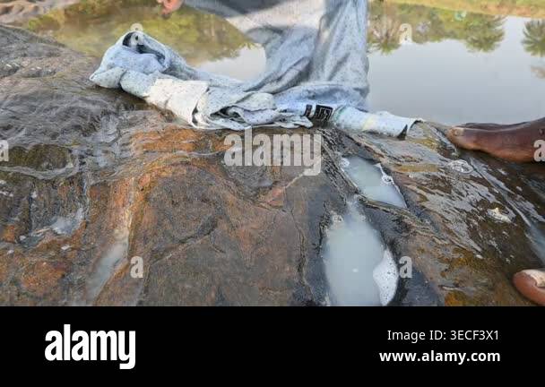Washing Clothes Using Rocks A man is washing clothes by hand on a stone at the riverbank. He uses ...