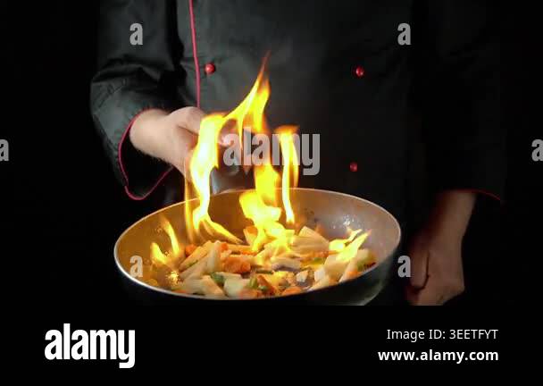 Chef in black uniform expertly flips vegetables in a flaming pan ...