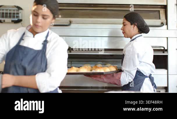 Diverse female bakers responding to oven timer, carrying metal trays to ...