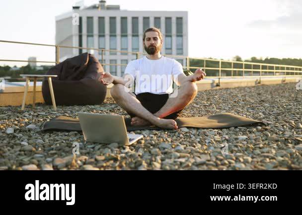 A man with a beard and long hair sits in a lotus position on a yoga mat ...