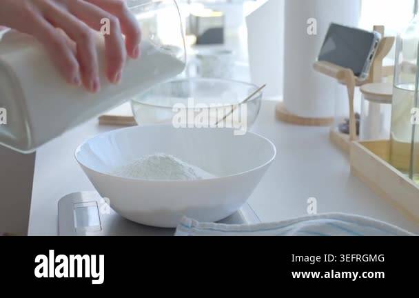 Hand pours flour from container into white bowl on kitchen scale ...