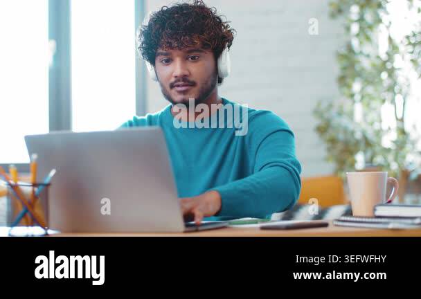 A young man with curly hair sits at a desk working on his laptop while ...