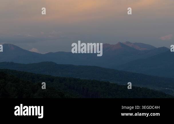 Moody sunset over forested slopes and layered mountain peaks fading ...