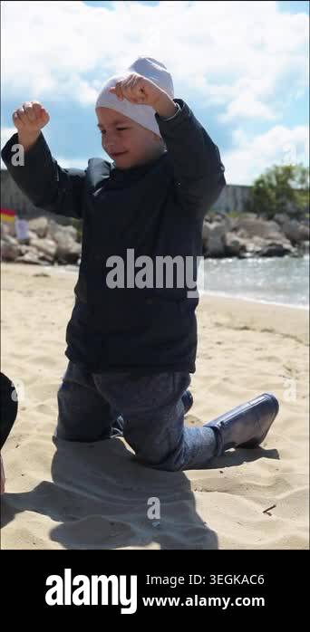 little boy in dark jacket and white hat kneeling on beach pouring sand ...