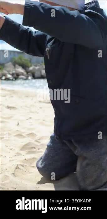 little boy in dark jacket and white hat kneeling on beach pouring sand ...