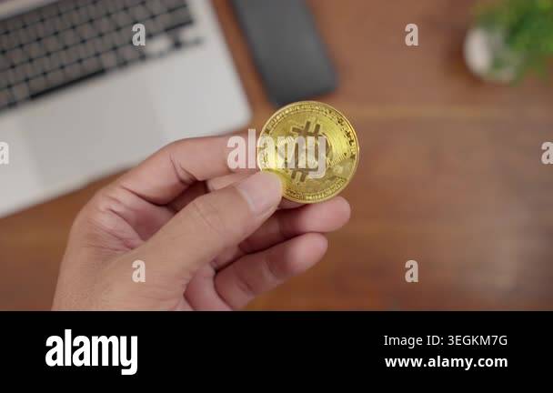 Close-up of a hand holding a gold Bitcoin coin on a work desk ...
