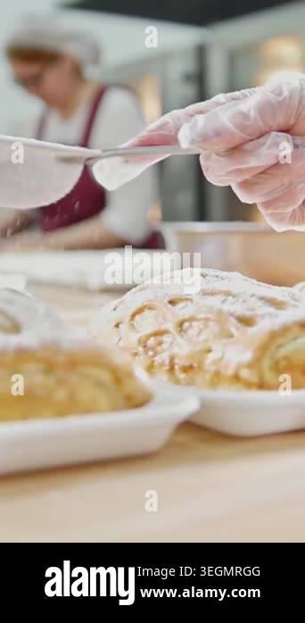 A baker uses a sieve to sprinkle sugar on a pastry in a busy kitchen ...