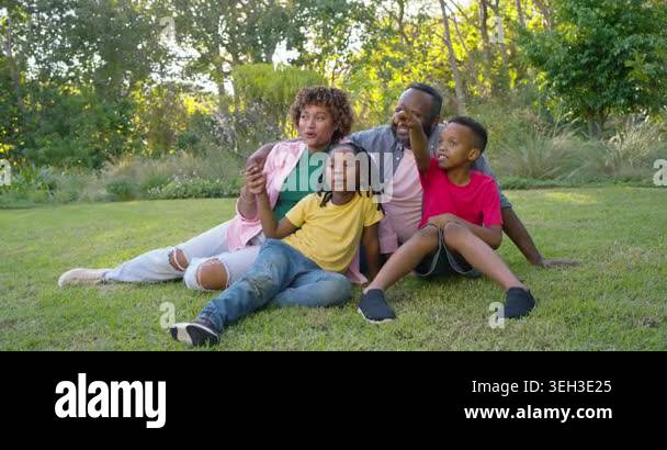 African American family sitting on lawn in park, child in red pointing ...