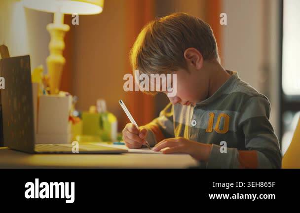 A young boy sits at a desk in a warm room with a laptop and lamp. He ...