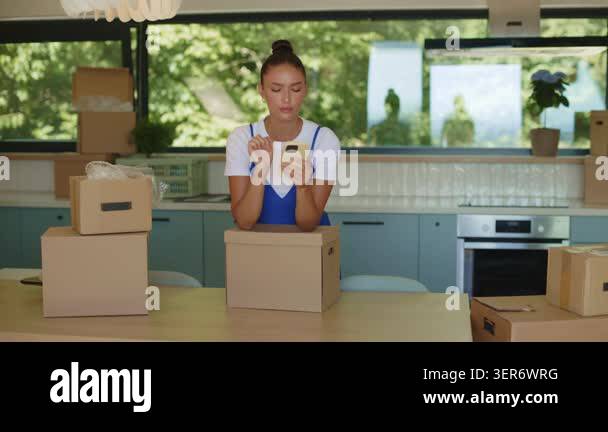 Young woman using smartphone while leaning on cardboard moving box in ...