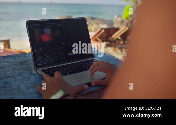 Male Video Editor Working on a Laptop at a Beach Cafe During Sunset ...