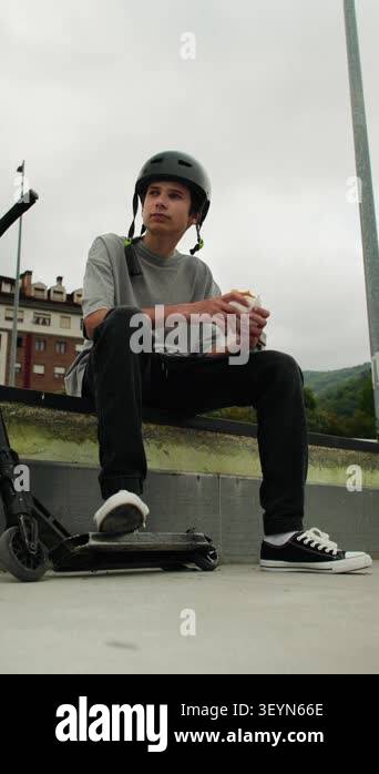 A teenage boy in a helmet sits at a skatepark next to his scooter ...