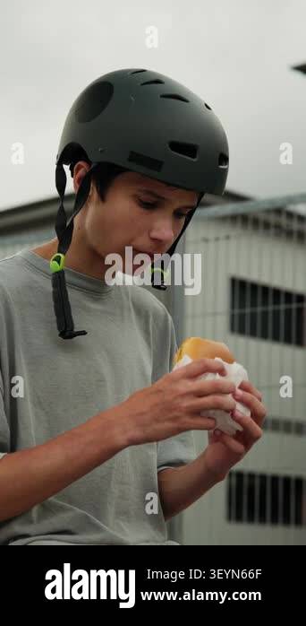 A teenage boy wearing a protective helmet eats a hamburger outdoors at ...