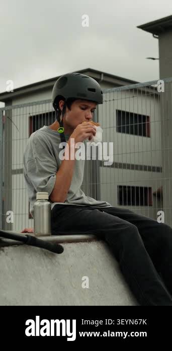 A teenage boy wearing a helmet sits on a ramp at a skatepark and eats a ...